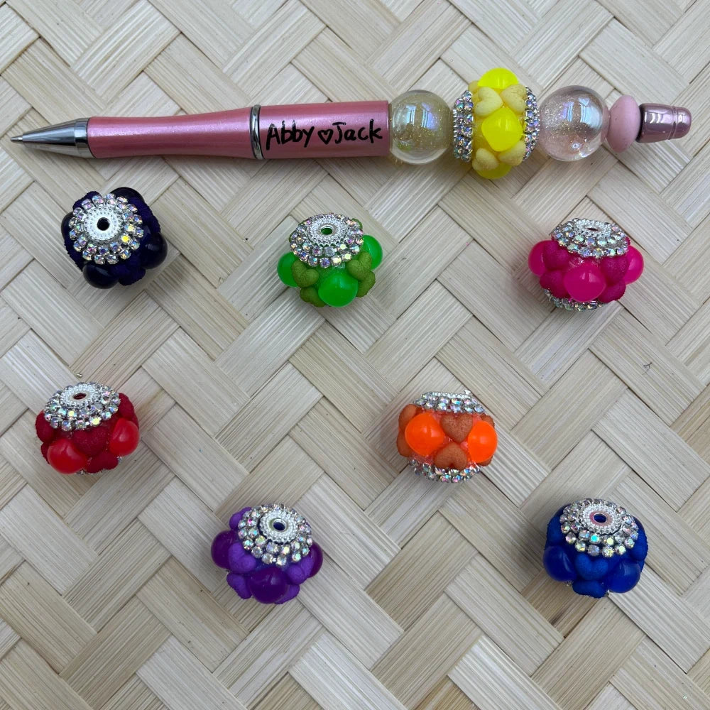Colorful rhinestone bubble cluster fancy beads in purple, green, pink, red, orange, blue, and yellow arranged next to a pink beadable pen labeled Abby and Jack on a woven rattan surface.