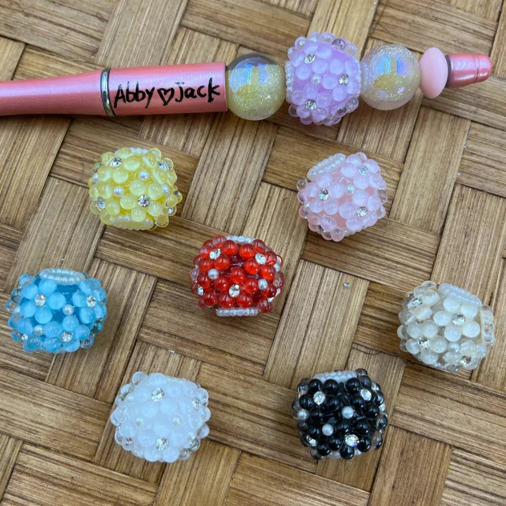 Assorted rhinestone bubble cluster fancy beads in yellow, red, blue, pink, white, black, and clear iridescent colors displayed on a wicker surface next to a pink beadable pen labeled Abby Jack.