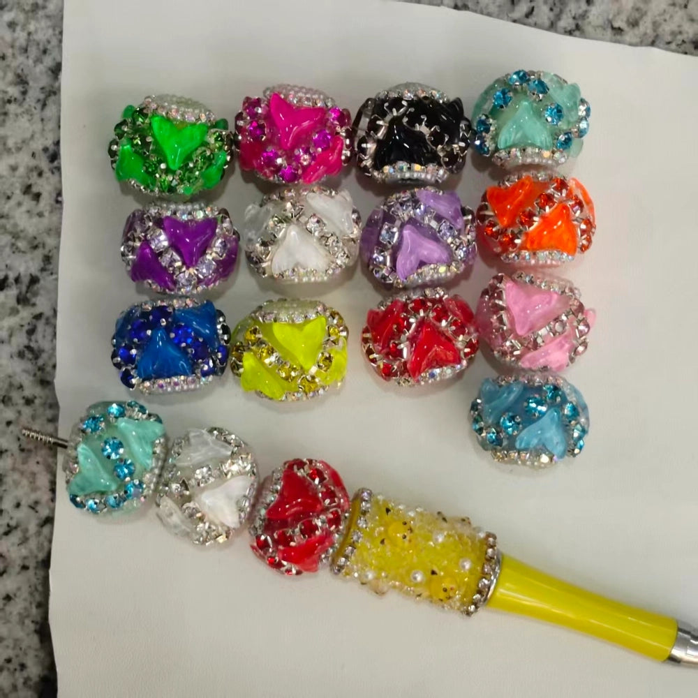 16 colorful rhinestone butterfly heart fancy beads in assorted colors including green, pink, black, teal, purple, white, lavender, orange, blue, yellow, red, and light pink, displayed on white paper with a beaded pen sample showing how beads look strung together.
