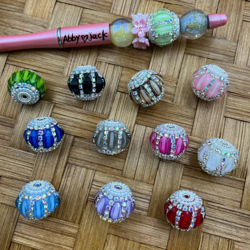 Assorted rhinestone cage fancy beads in multiple colors including green, black, blue, pink, red, purple, white, and gray with crystal AB rhinestone and pearl trim accents, displayed on wicker background next to a decorated beadable pen.