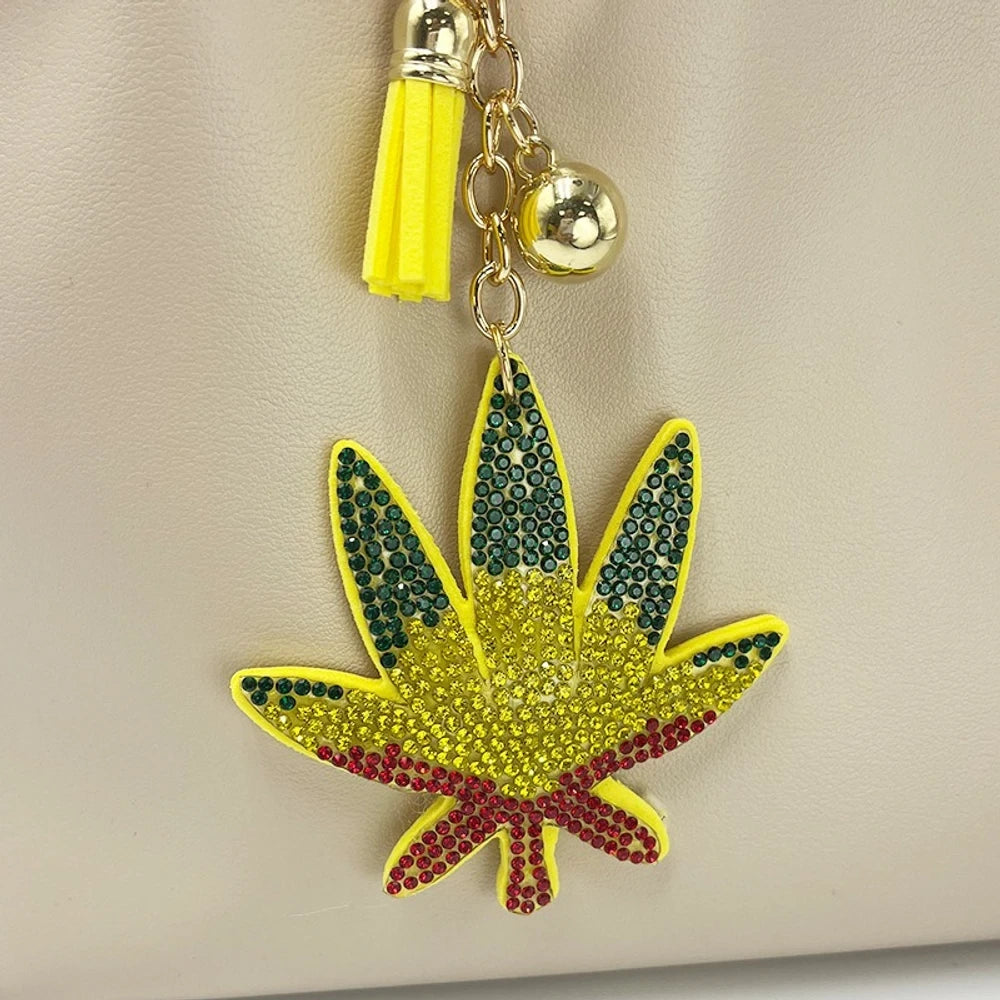 Rhinestone cannabis marijuana leaf keychain charm with yellow tassel and gold chain, decorated with green, yellow, and red crystal rhinestones on a cream leather bag. - Green