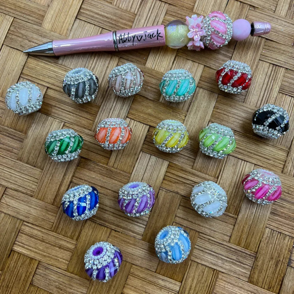 Assorted rhinestone-bordered cat eye stripe fancy beads in multiple colors including pink, teal, red, green, orange, yellow, purple, blue, black, and white, displayed on a woven surface next to a decorated beadable pen.