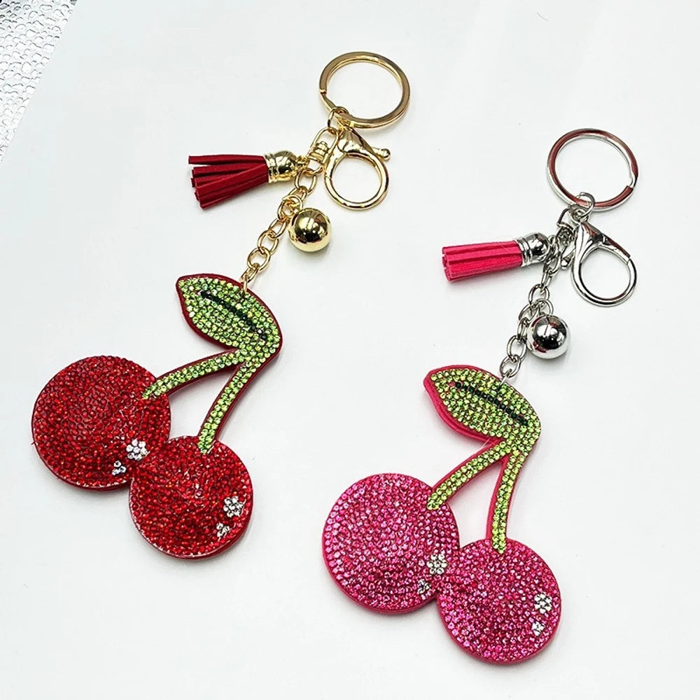Two rhinestone-encrusted cherry keychain charms with green leaf crystals, tassels, and bells — one red on gold hardware, one pink on silver hardware.