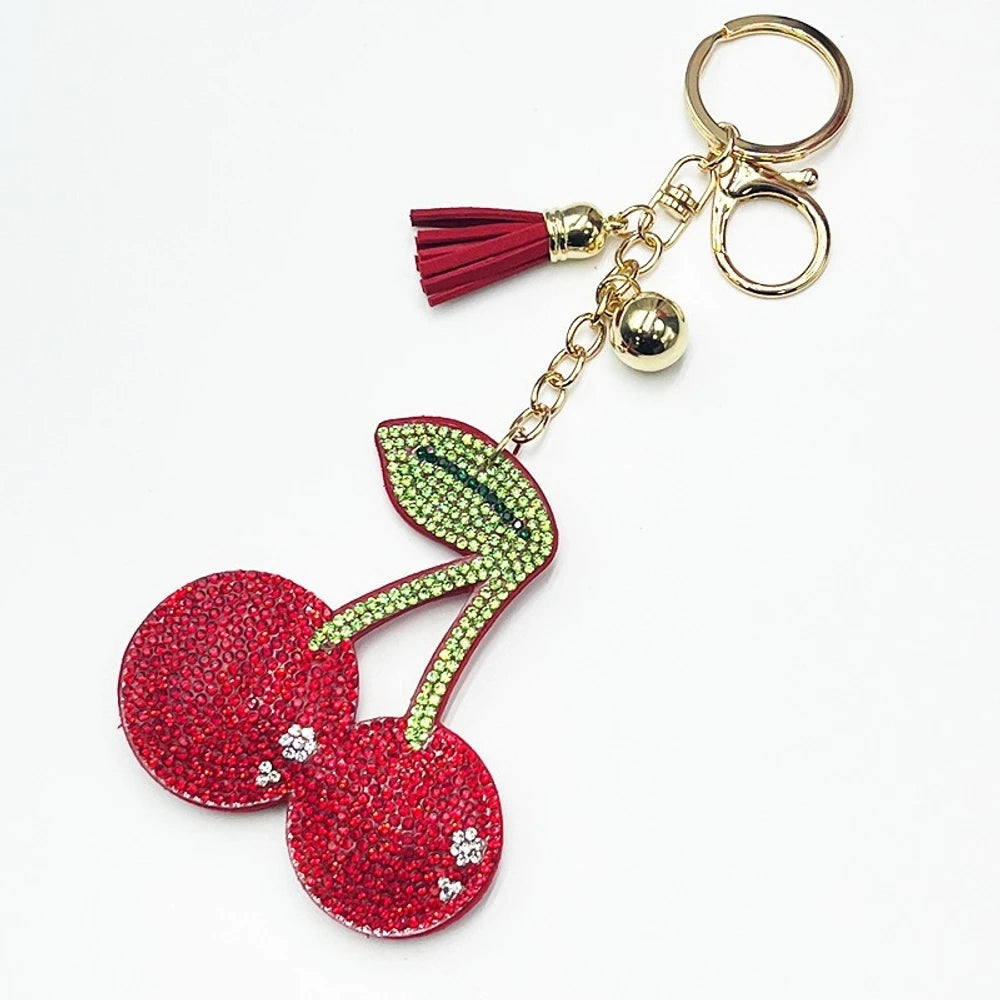 Two rhinestone-encrusted cherry keychain charms with green leaf crystals, tassels, and bells — one red on gold hardware, one pink on silver hardware. - Red