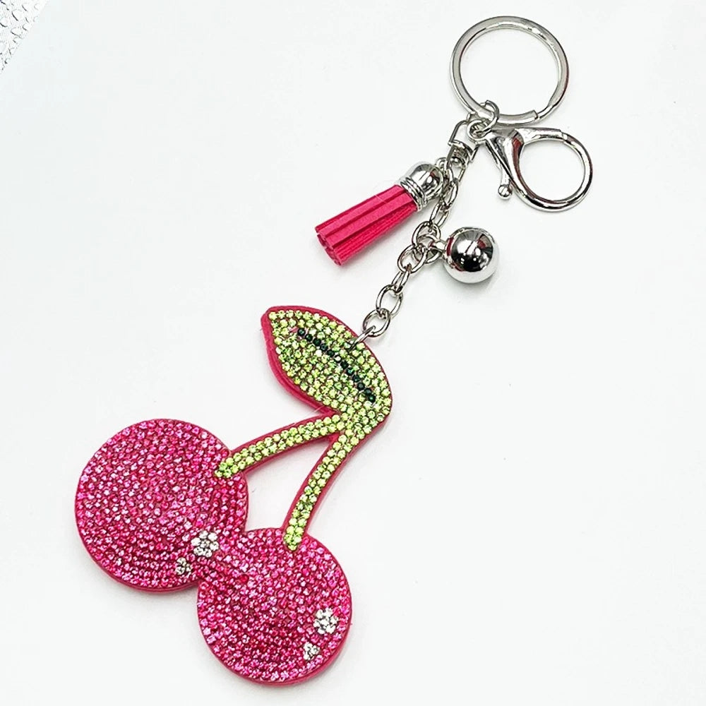 Two rhinestone-encrusted cherry keychain charms with green leaf crystals, tassels, and bells — one red on gold hardware, one pink on silver hardware. - Rose Pink