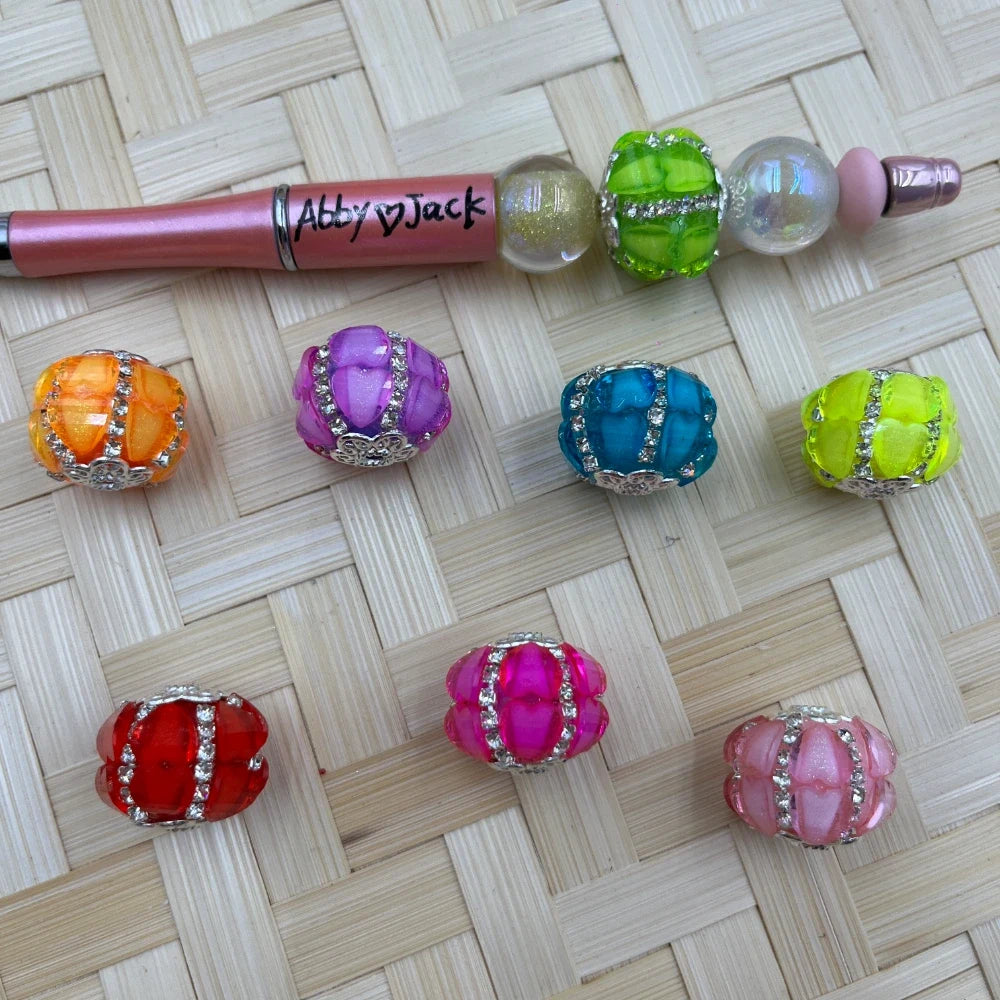 Colorful rhinestone studded chunky resin fancy beads in orange, purple, teal, yellow-green, red, hot pink, and light pink, displayed next to a beadable pen on a wicker surface.
