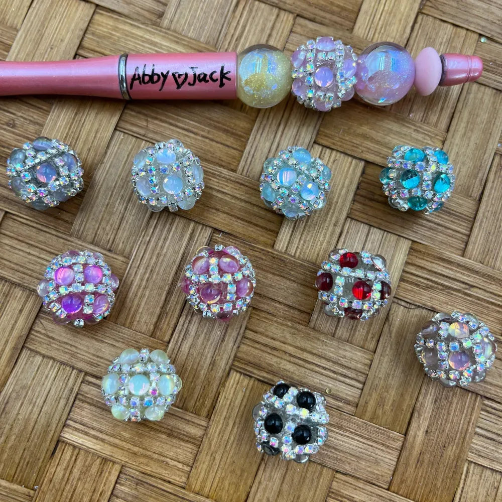 Assorted rhinestone-rimmed cluster fancy beads in multiple colors including clear, pink, teal, red, black, and iridescent opal, displayed on a wicker surface next to a decorated beadable pen labeled Abby Jack.