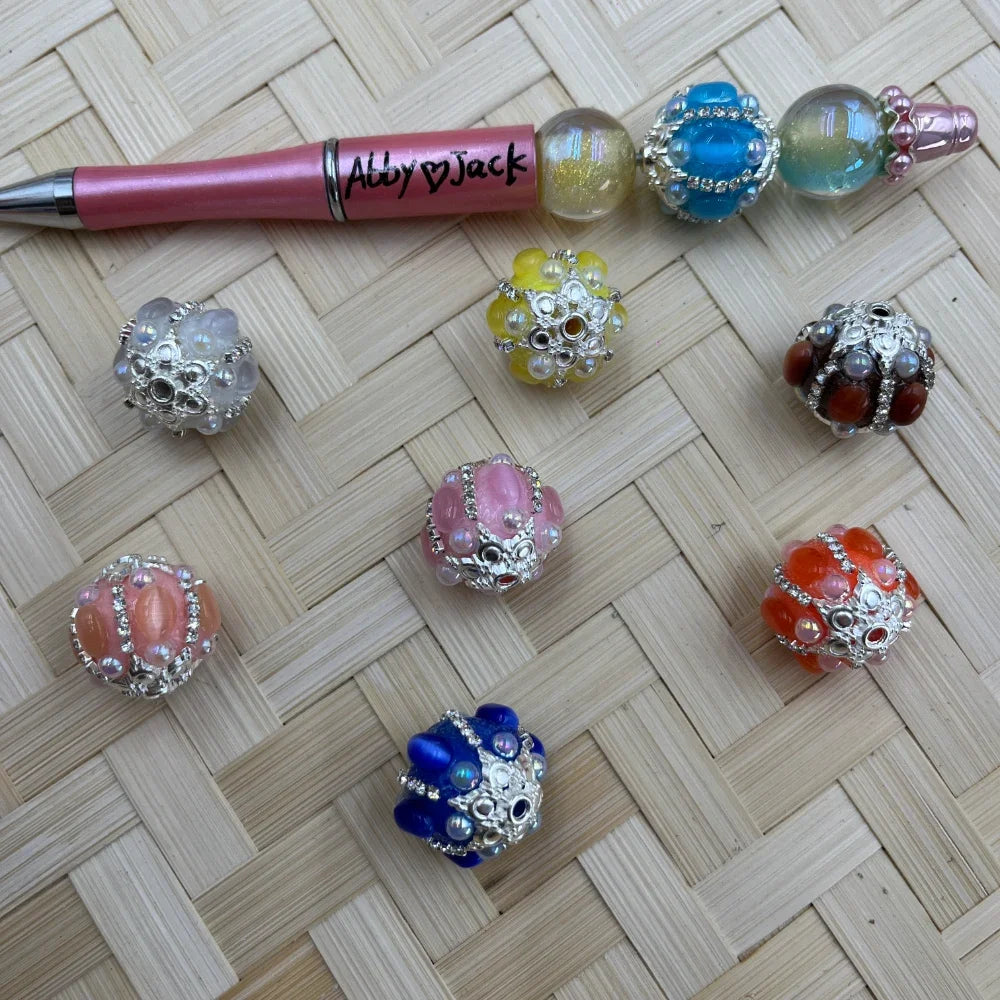 Assorted rhinestone cluster fancy beads in pink, orange, blue, yellow, white, and red-brown with silver metal filigree caps, displayed next to a pink beadable pen on a woven bamboo background.