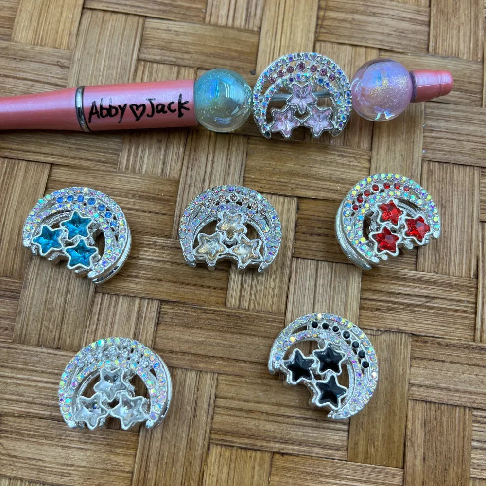 Assorted rhinestone crescent moon and star metal beads in blue, red, clear, black, and pink displayed on a woven surface next to a pink beadable pen labeled Abby and Jack.