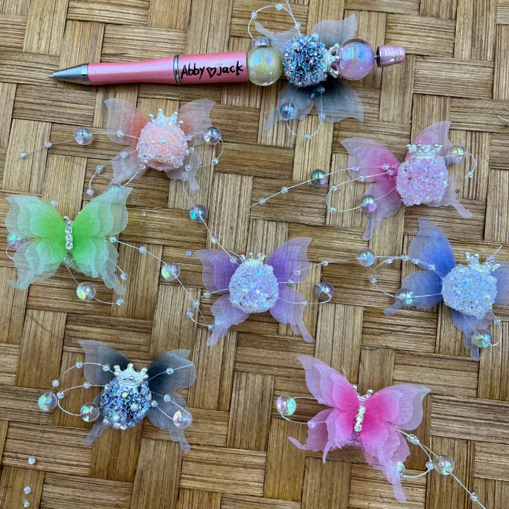 Assorted rhinestone crown butterfly fancy beads with sheer organza wings and iridescent pearl wire accents in pink, purple, green, blue, and gray, displayed alongside a decorated beadable pen on a wicker surface.