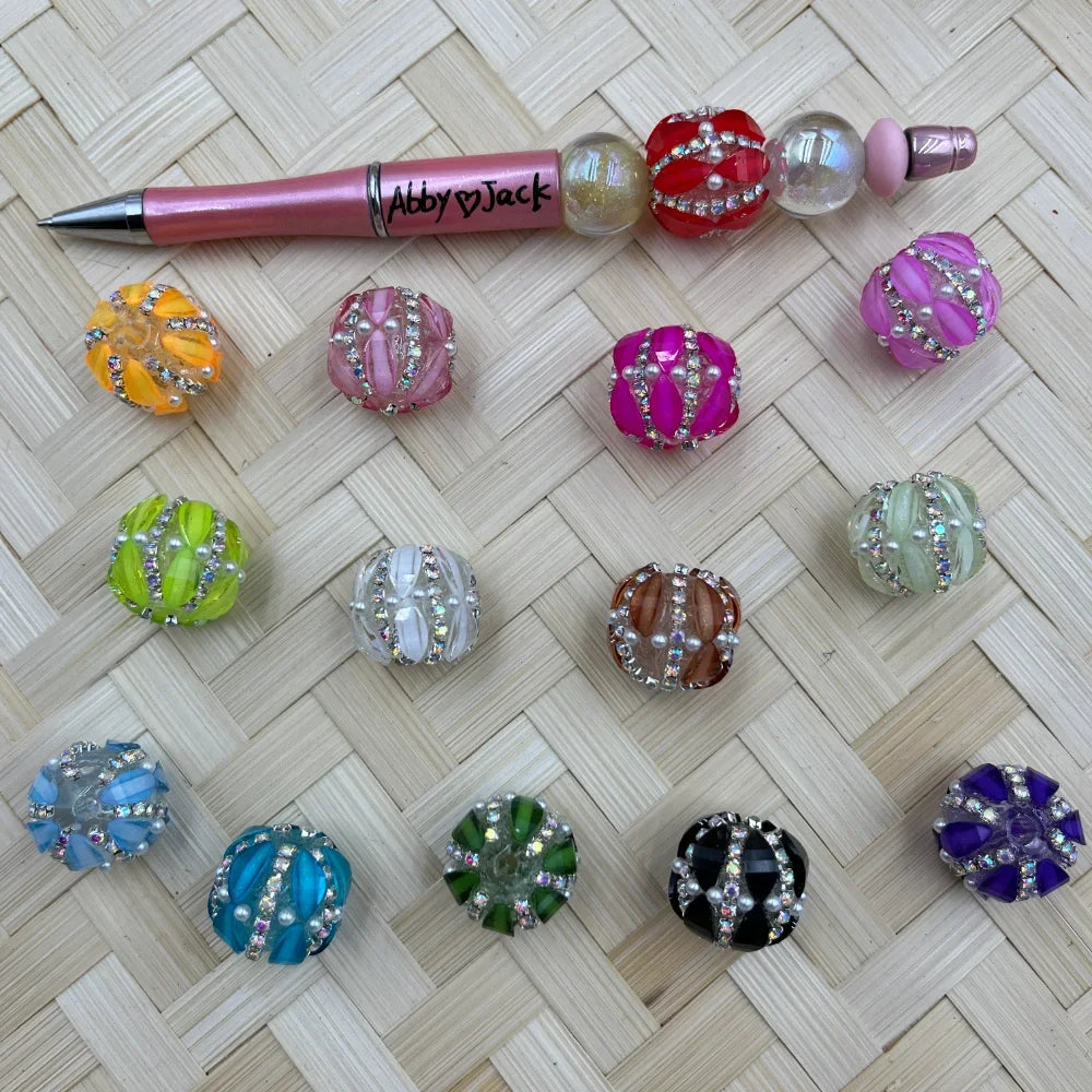 Assorted rhinestone crown fancy beads in multiple colors including pink, orange, green, blue, purple, black, white, and red displayed on a woven surface next to a beadable pen labeled Abby Jack.