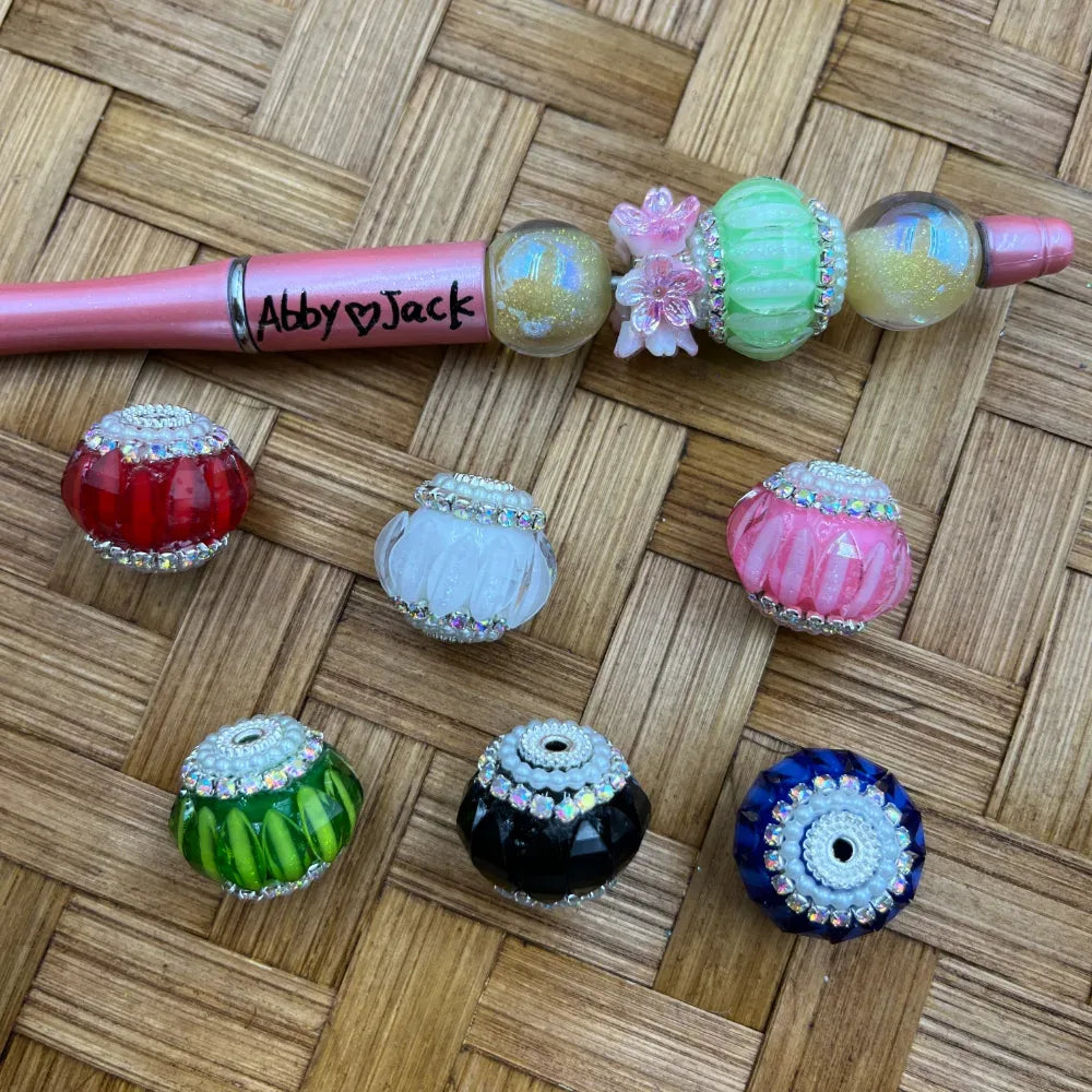 Assorted rhinestone crown fancy beads in red, white, pink, green, black, and blue with silver crystal trim, displayed alongside a decorated pink beadable pen on a woven bamboo surface.