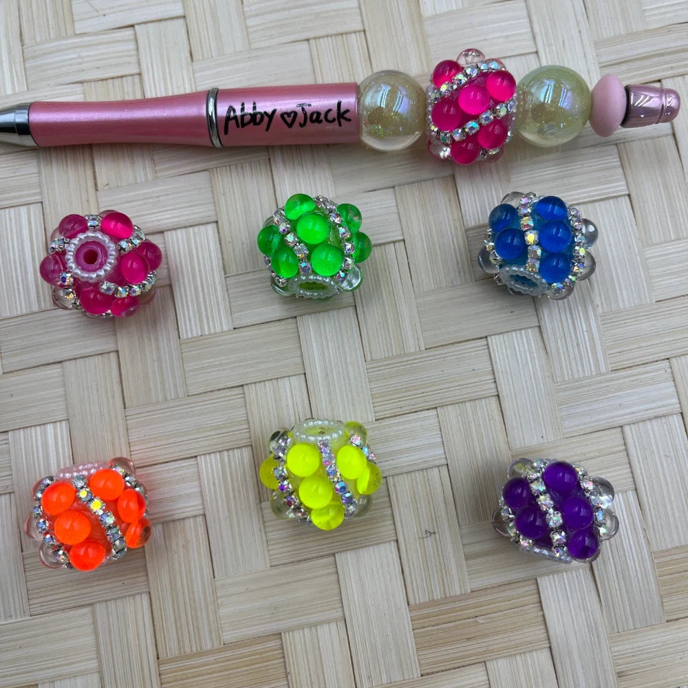 Assorted neon rhinestone crystal cluster fancy beads in pink, green, blue, orange, yellow, and purple displayed on a woven surface next to a beadable pen labeled Abby Jack, for DIY jewelry making and beadable pens.