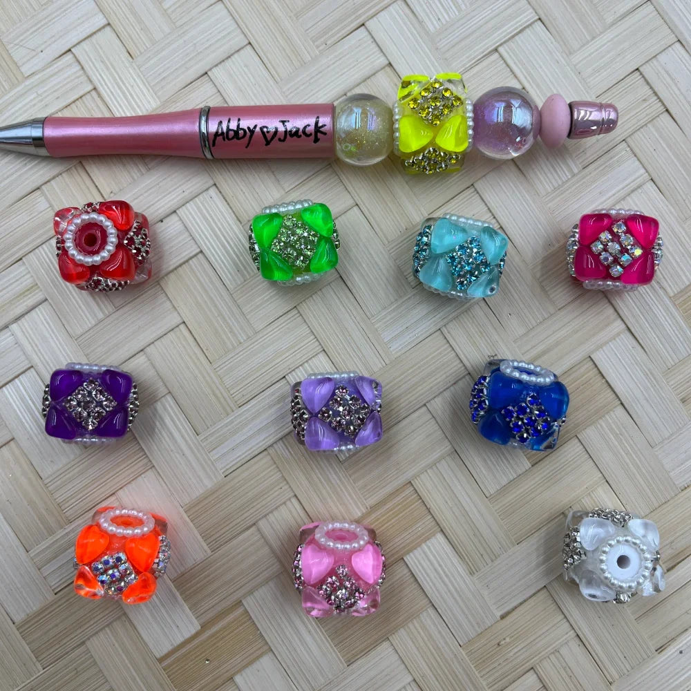 Assorted colorful rhinestone crystal cube fancy beads in red, green, teal, pink, purple, lavender, blue, orange, and clear with pearl and gemstone accents, displayed on a woven surface next to a decorated beadable pen labeled Abby and Jack.