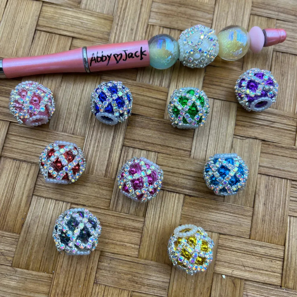Assorted rhinestone crystal diamond pattern fancy beads in pink, blue, green, purple, red, magenta, teal, grey, and yellow with iridescent AB finish, displayed next to a beadable pen on a wicker surface.