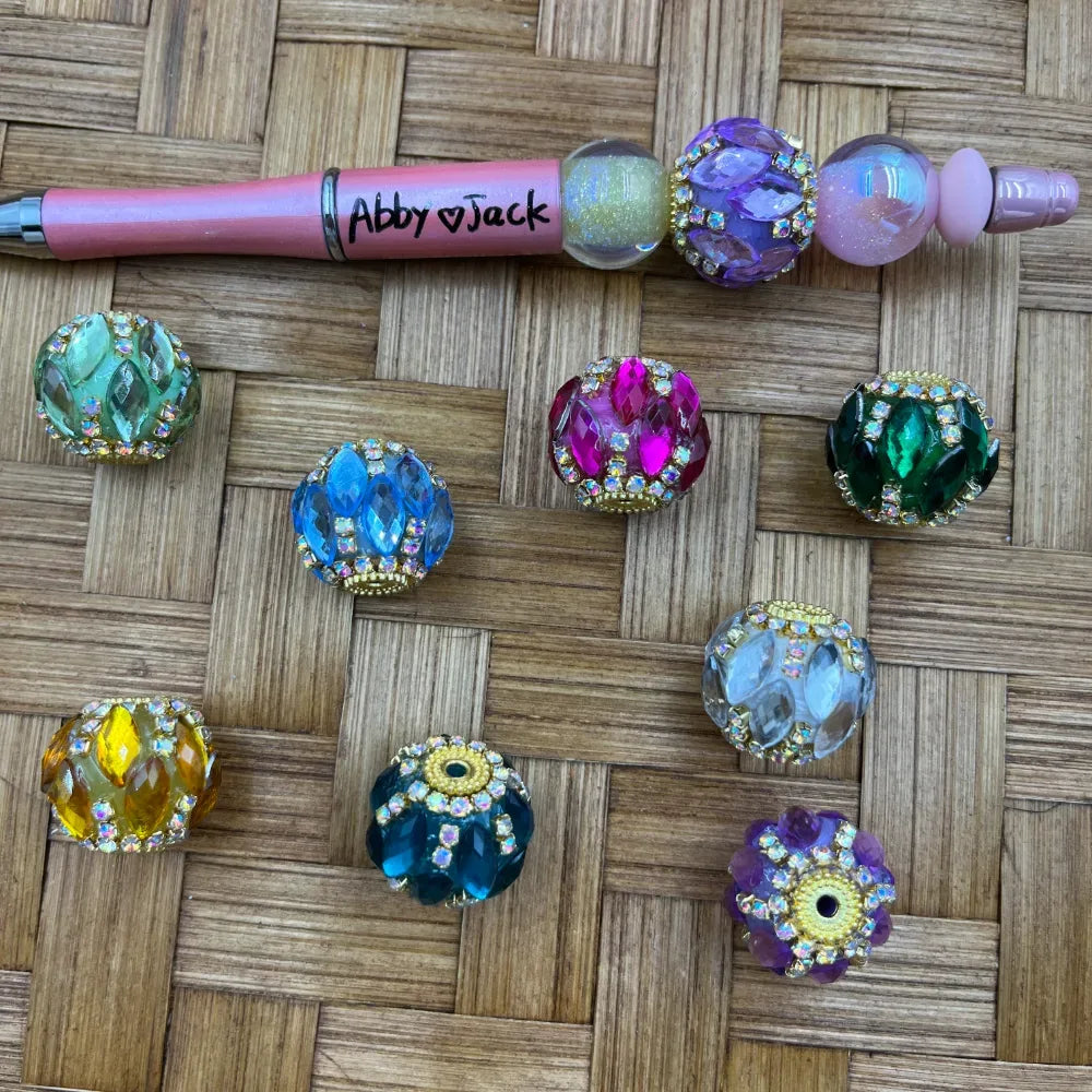 Assorted rhinestone-trimmed crystal fancy beads in green, blue, pink, dark green, yellow, teal, clear, and purple colors displayed on woven surface next to a pink beadable pen labeled Abby and Jack.