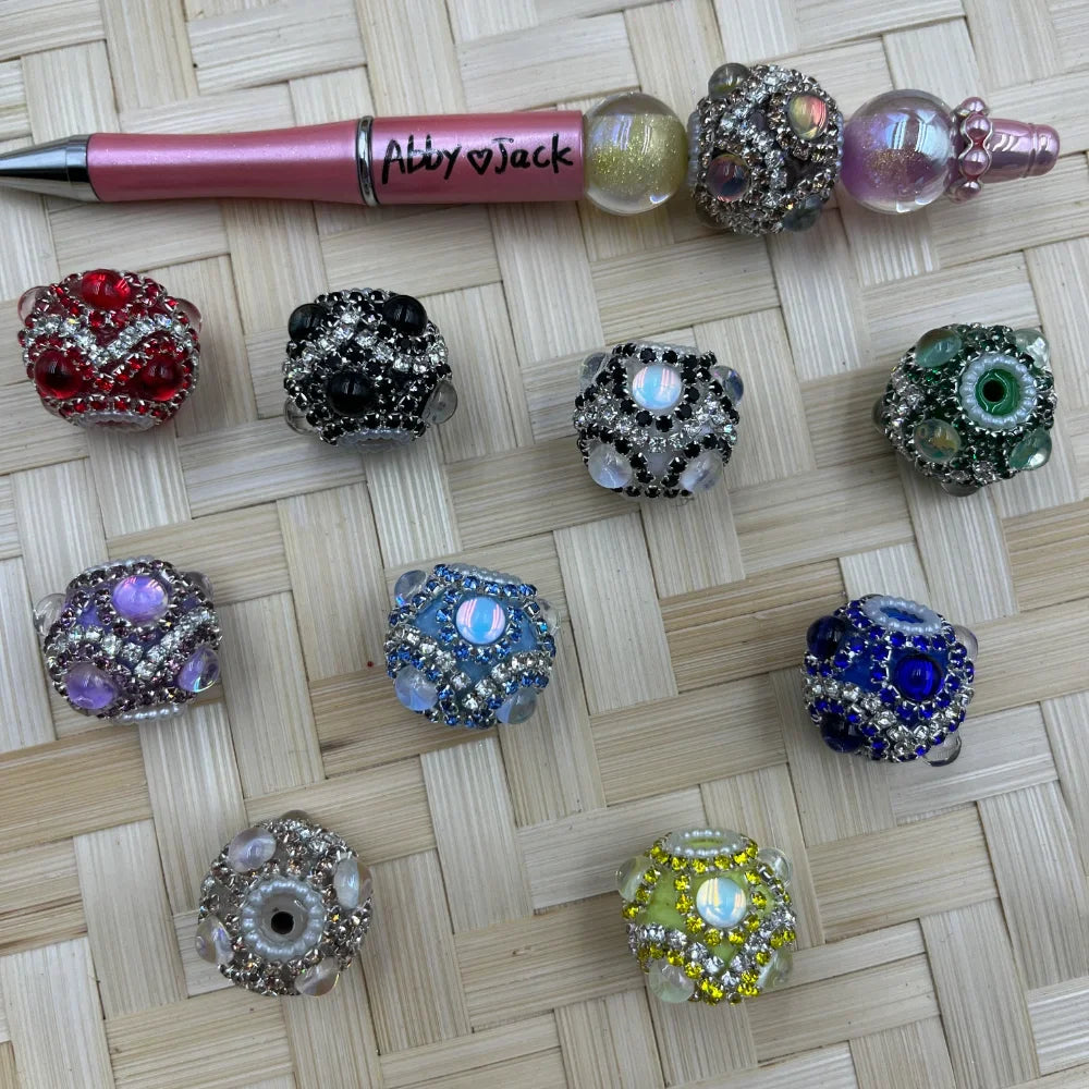 Assorted rhinestone crystal fancy beads in red, black, purple, blue, green, yellow and clear displayed on woven surface next to a beadable pen from DIY Mini Planet