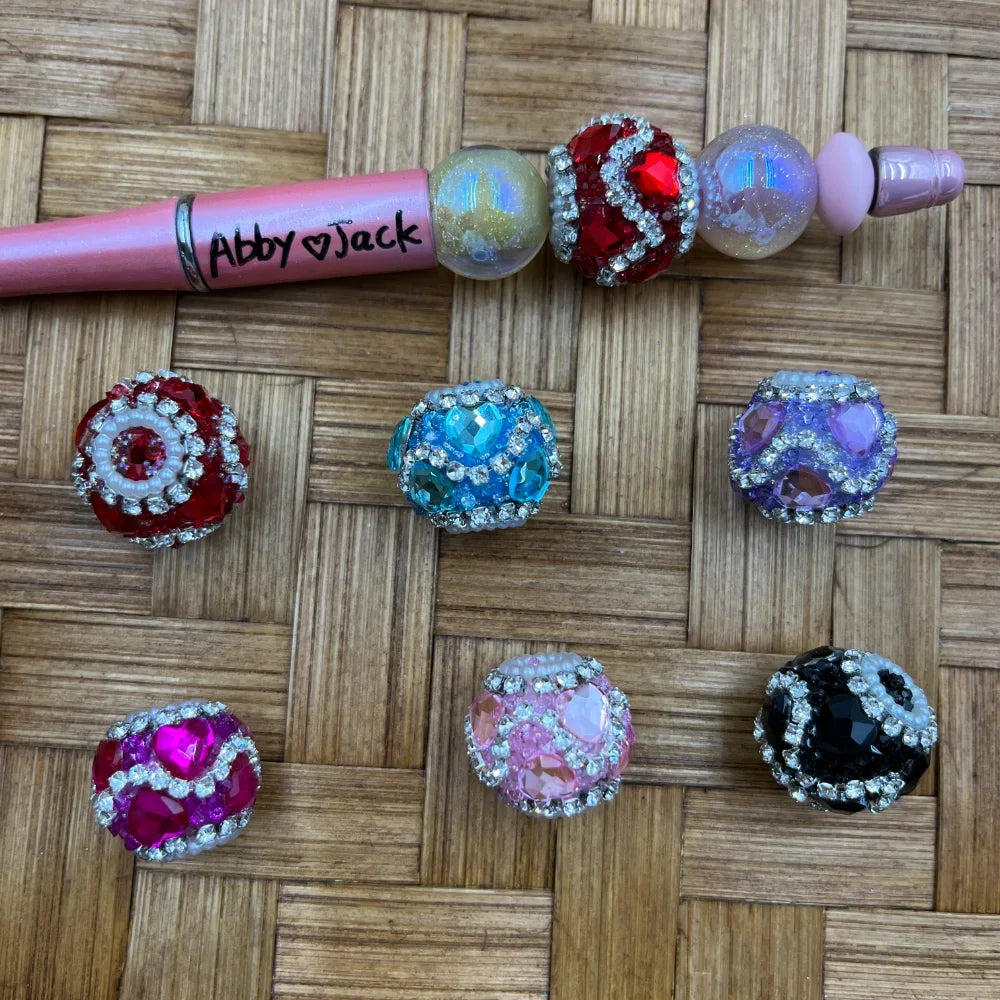 Assorted rhinestone crystal fancy beads in red, turquoise, purple, hot pink, light pink, and black with silver diamond-cut accents displayed on a woven surface next to a pink beadable pen labeled Abby and Jack.