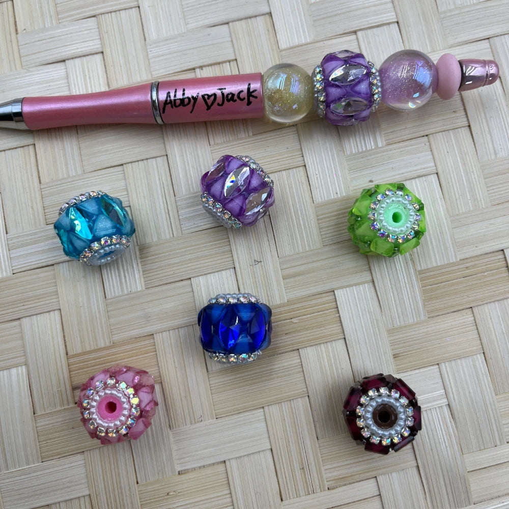 Assorted rhinestone crystal fancy beads in teal, purple, green, blue, pink, and burgundy displayed on woven surface next to a beaded pink pen labeled Abby Jack.