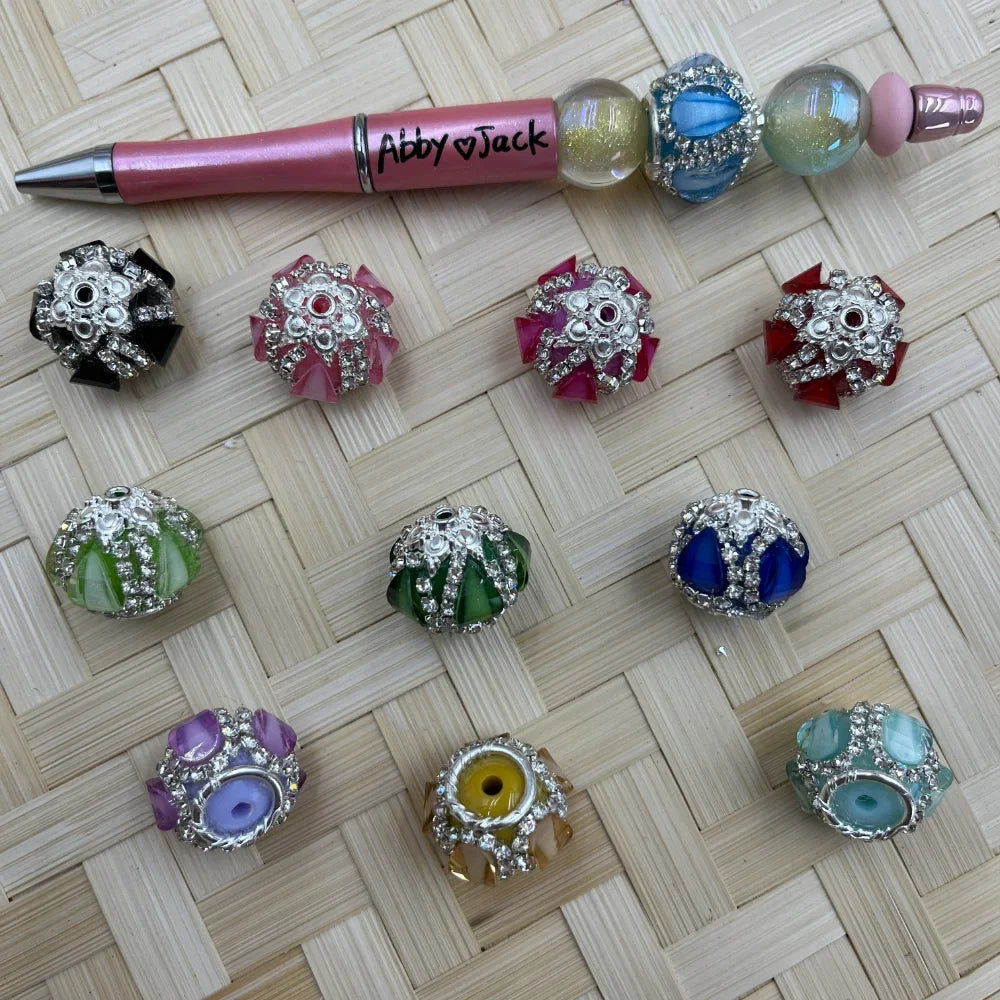 Assorted rhinestone crystal fancy beads in black, pink, purple, magenta, red, light green, dark green, blue, lavender, yellow, and teal displayed on woven surface alongside a pink beadable pen labeled Abby and Jack