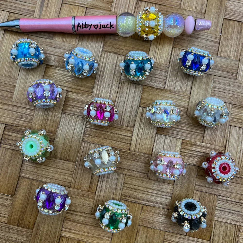 Assorted rhinestone crystal fancy beads in multiple colors including blue, teal, purple, pink, green, red, and black with pearl and gemstone accents, displayed on a woven surface next to a pink beadable pen labeled Abby Jack.