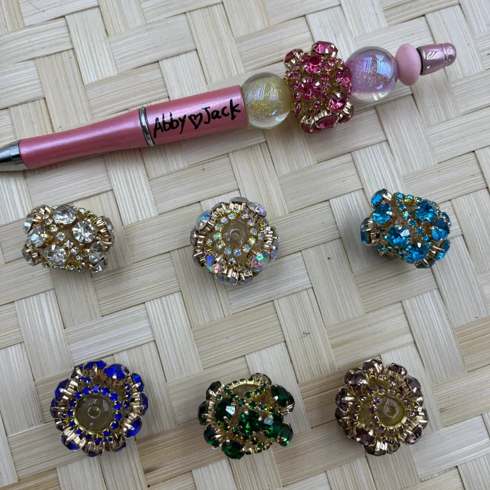 Assorted crystal rhinestone flower fancy beads in clear, aurora borealis, teal, blue, green, and purple with gold-tone settings, displayed with a pink beadable pen on wicker surface.