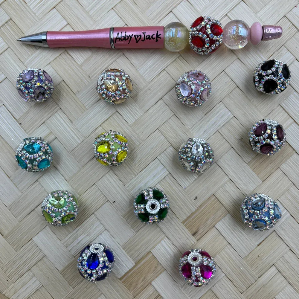 Assorted rhinestone crystal gem fancy beads in multiple colors including purple, teal, yellow, green, blue, pink, red, and black displayed on a woven surface next to a pink beadable pen labeled Abby and Jack