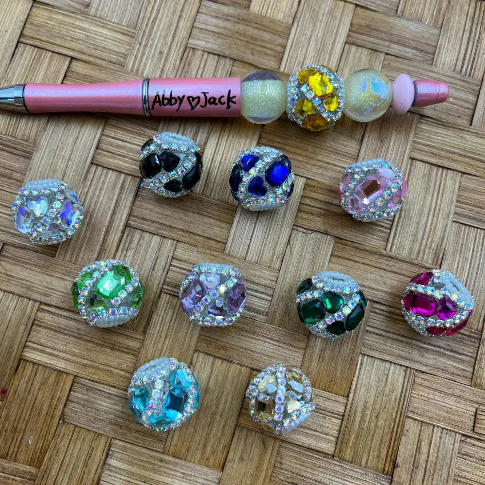 Assorted rhinestone crystal gem fancy beads in multiple colors including black, blue, pink, green, teal, lavender, magenta, and yellow, displayed on a woven surface alongside a pink beadable pen labeled Abby and Jack