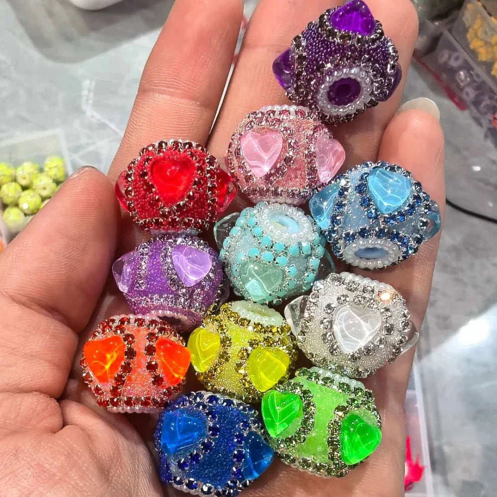 Hand holding 14 colorful rhinestone-encrusted fancy beads each featuring a translucent crystal heart center in colors including red, pink, purple, blue, green, yellow, orange, teal, and clear