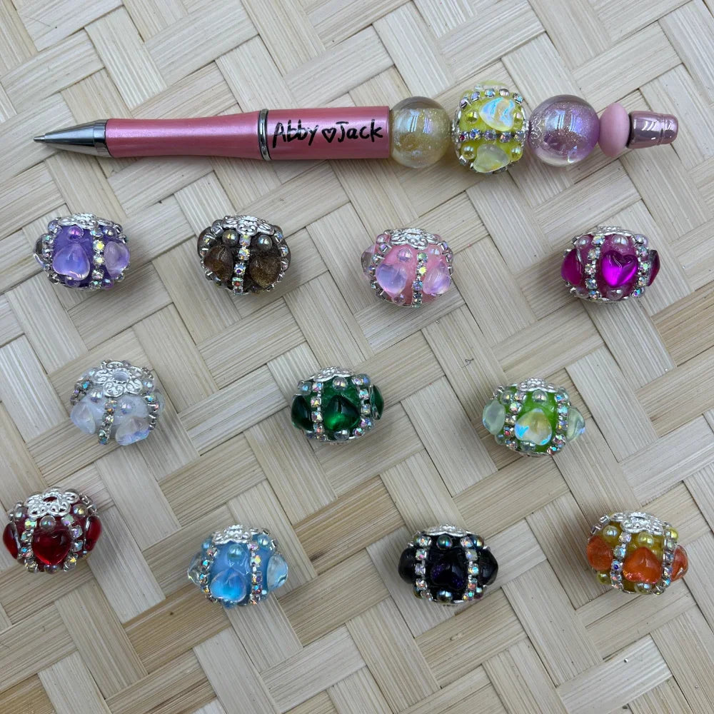 Assorted rhinestone crystal heart fancy beads in 11 colors including purple, green, pink, black, red, blue, and orange with silver filigree caps and AB rhinestone accents, displayed next to a pink beadable pen on a woven surface.