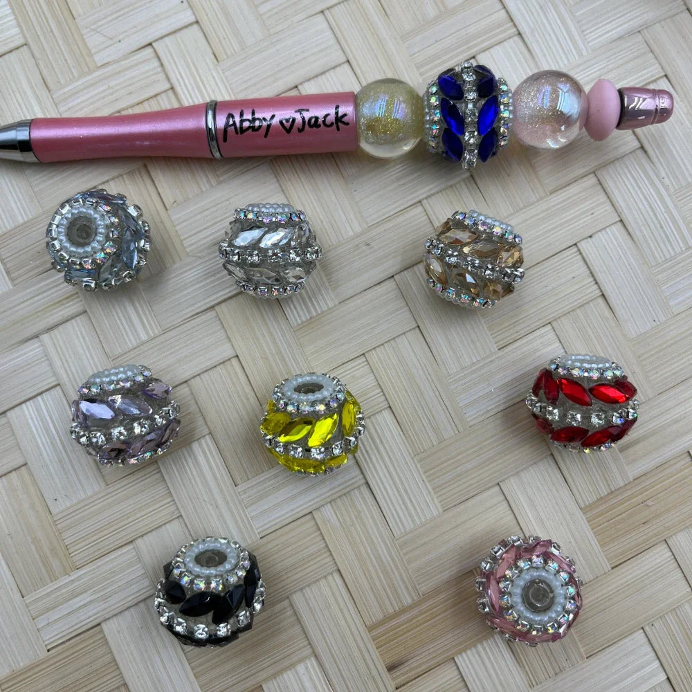 Assorted rhinestone crystal rondelle fancy beads in multiple colors including clear, blue, champagne, lavender, yellow, red, black, and pink, displayed next to a decorated beadable pen on a wicker surface.