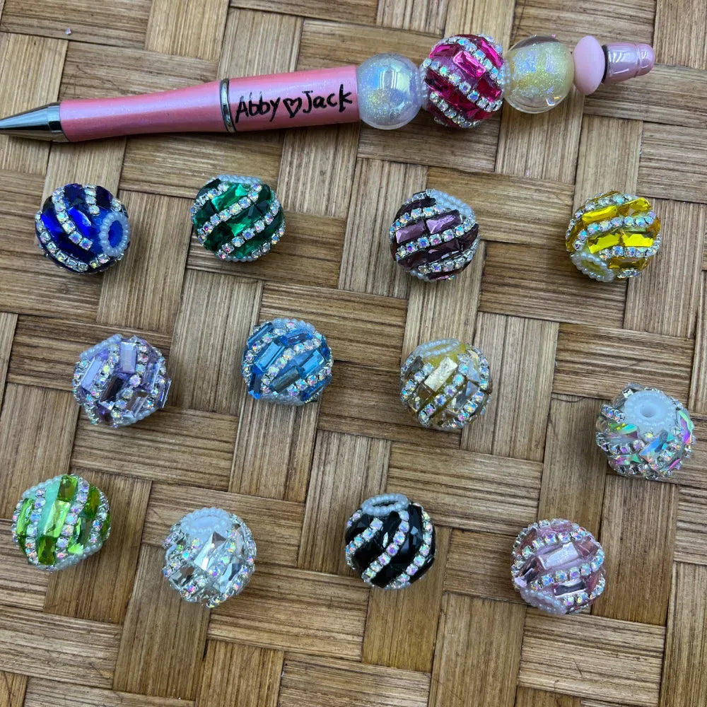 Assorted rhinestone crystal striped chunky round fancy beads in multiple colors including blue, green, purple, yellow, pink, black, and clear, displayed on a woven surface next to a pink beadable pen labeled Abby and Jack.