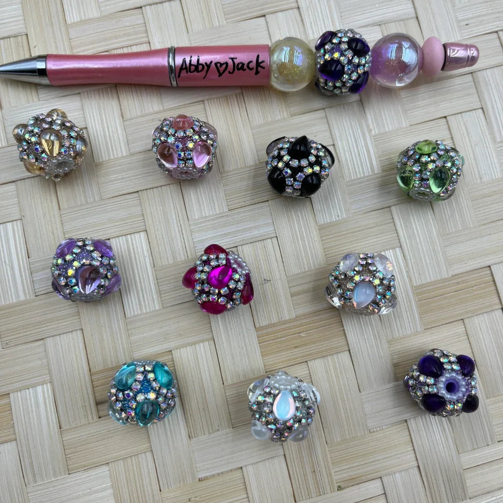 Assorted rhinestone crystal teardrop fancy beads in purple, pink, teal, black, green, and clear on woven surface next to a pink beadable pen labeled Abby Jack