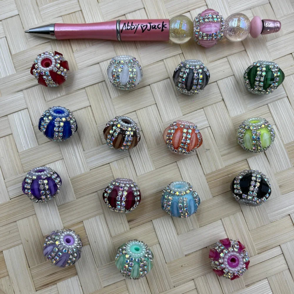 Assorted rhinestone encrusted fancy beads in multiple colors including red, white, black, green, blue, brown, orange, lime, purple, burgundy, teal, black, lavender, mint, and pink displayed on a woven surface alongside a pink beadable pen labeled Abby and Jack.