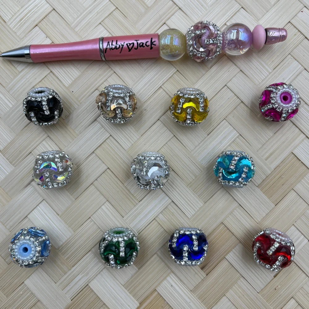 Assorted rhinestone encrusted swirl glass fancy beads in 12 colors including black, yellow, teal, green, blue, red, pink, and clear, displayed on woven surface next to a decorative beadable pen.