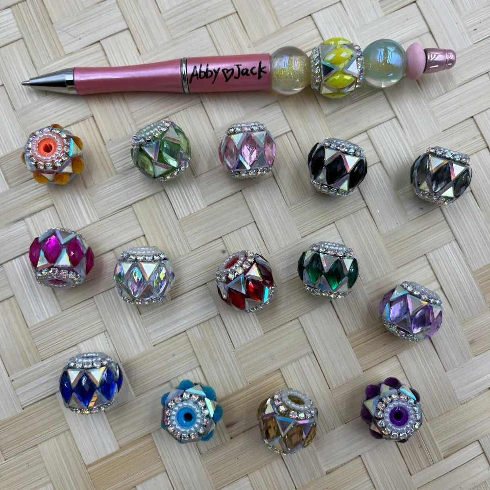 Assorted rhinestone-rimmed faceted gem fancy beads in multiple colors including green, pink, black, red, purple, blue, and iridescent AB finish, displayed on a woven bamboo surface next to a pink beadable pen labeled Abby and Jack.