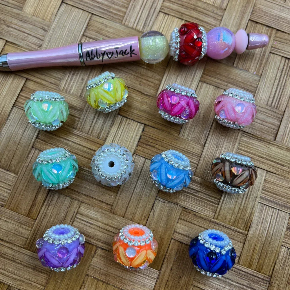 Assorted rhinestone-trimmed floral acrylic fancy beads in 12 colors including green, yellow, pink, teal, blue, purple, orange, and brown displayed on a woven surface next to a decorative pink beadable pen branded Abby Jack.