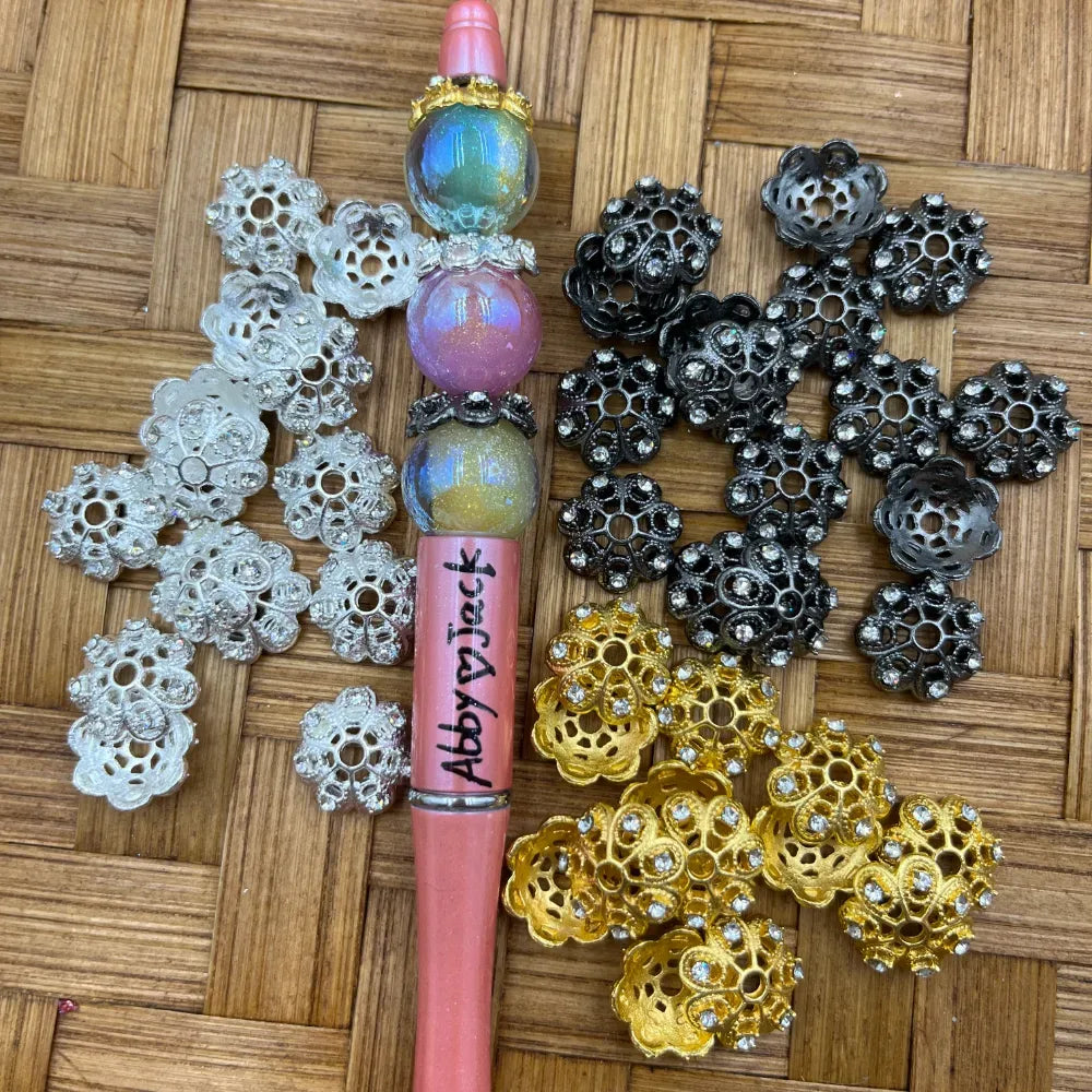 Assorted rhinestone flower bead caps in silver, black gunmetal, and gold finishes displayed next to a decorated beadable pen with iridescent beads on a wicker surface.