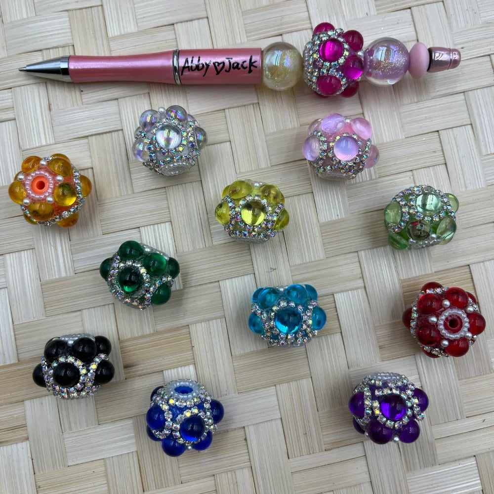 Assorted rhinestone flower bubble fancy beads in 11 colors including pink, green, blue, purple, red, black, yellow, and clear, displayed on a woven surface next to a beaded pen.