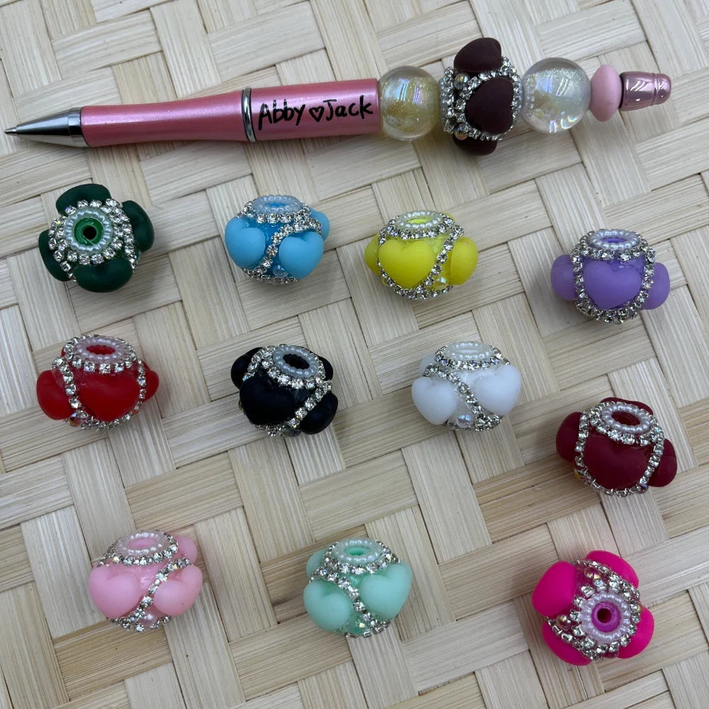 Assorted rhinestone-trimmed bubbly flower fancy beads in 11 colors including pink, red, black, white, teal, lavender, yellow, blue, green, dark red, and hot pink, displayed on a woven surface next to a beaded pen labeled Abby Jack.