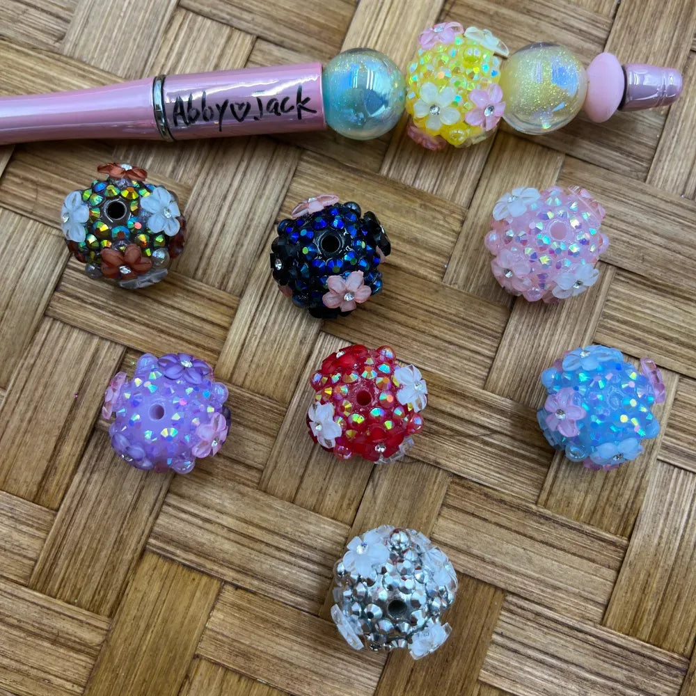 Assorted rhinestone and flower embellished chunky round beads in black, red, blue, pink, purple, white, and iridescent colors displayed on a wicker surface next to a pink beadable pen branded Abby Jack.