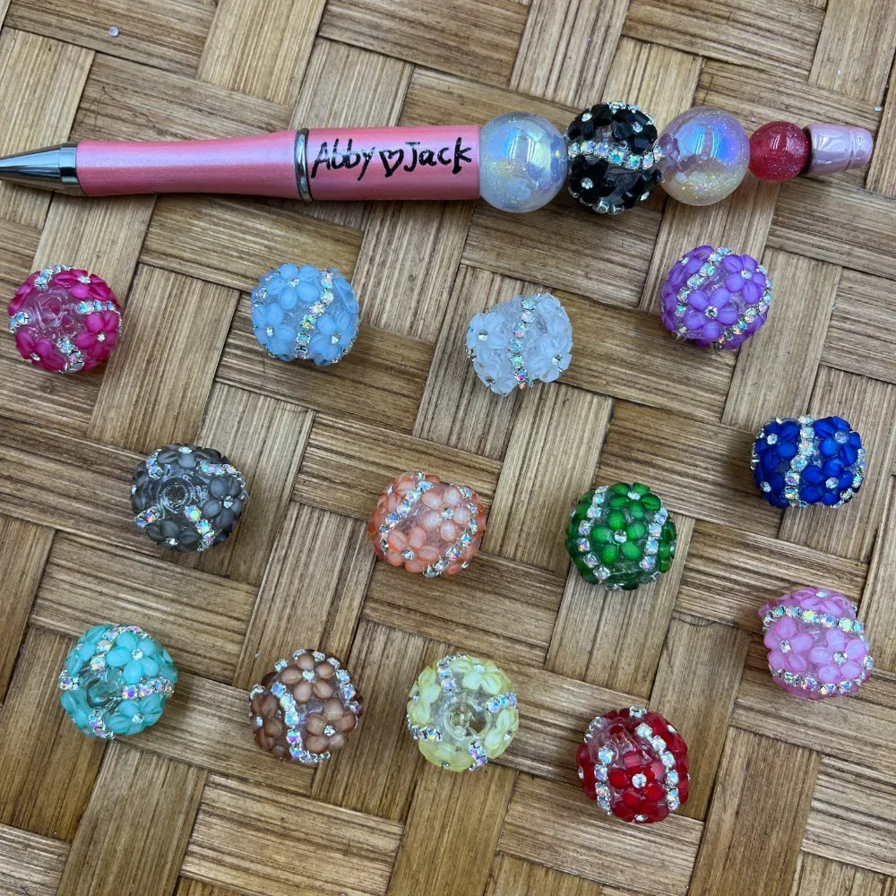 Assorted rhinestone-accented flower cluster acrylic fancy beads in 14 colors including pink, blue, green, red, purple, teal, gray, orange, and clear, displayed next to a decorative beadable pen on a wicker background.