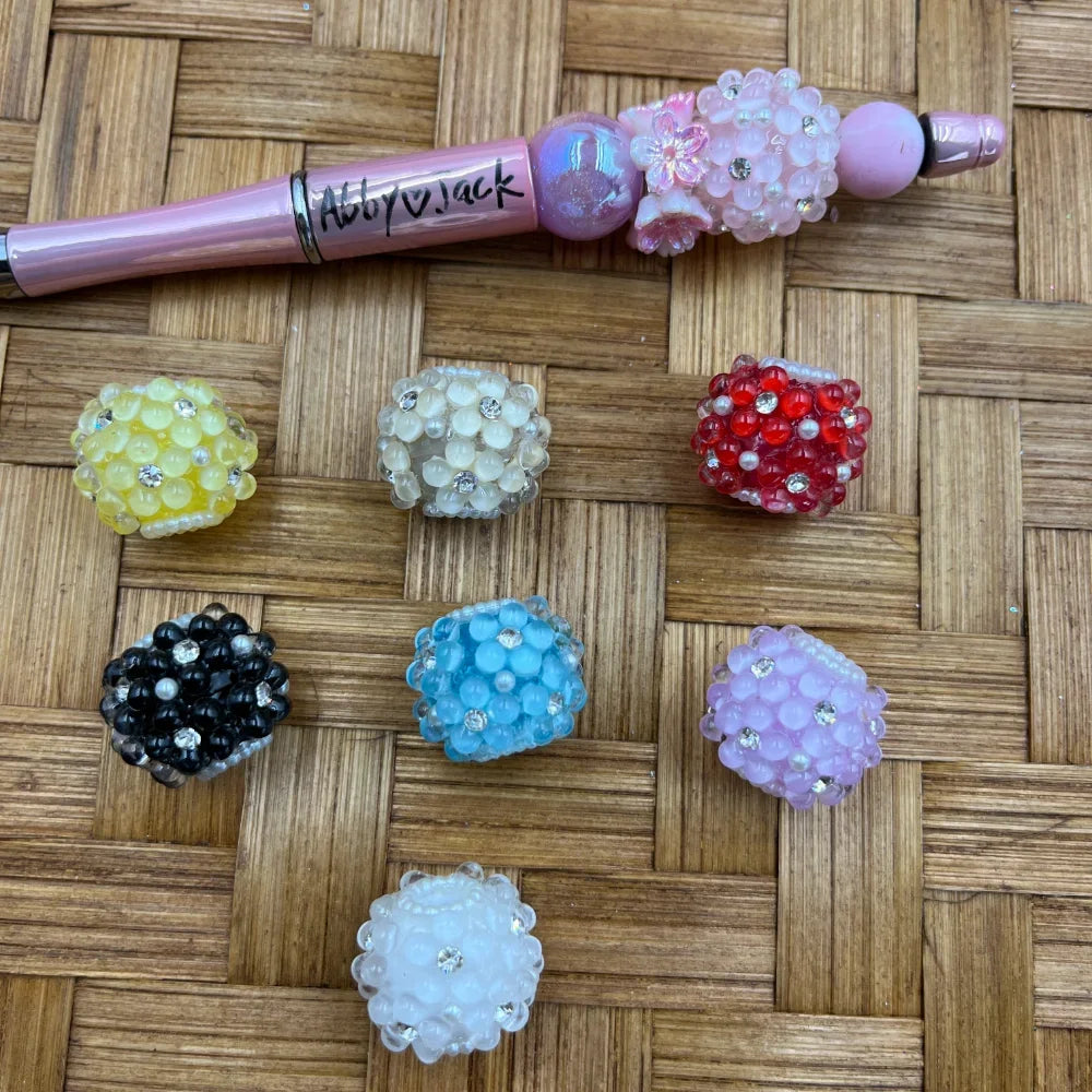 Assorted rhinestone flower cluster acrylic fancy beads in yellow, white, red, black, blue, lavender, and pink displayed on a woven surface next to a decorated beadable pen.