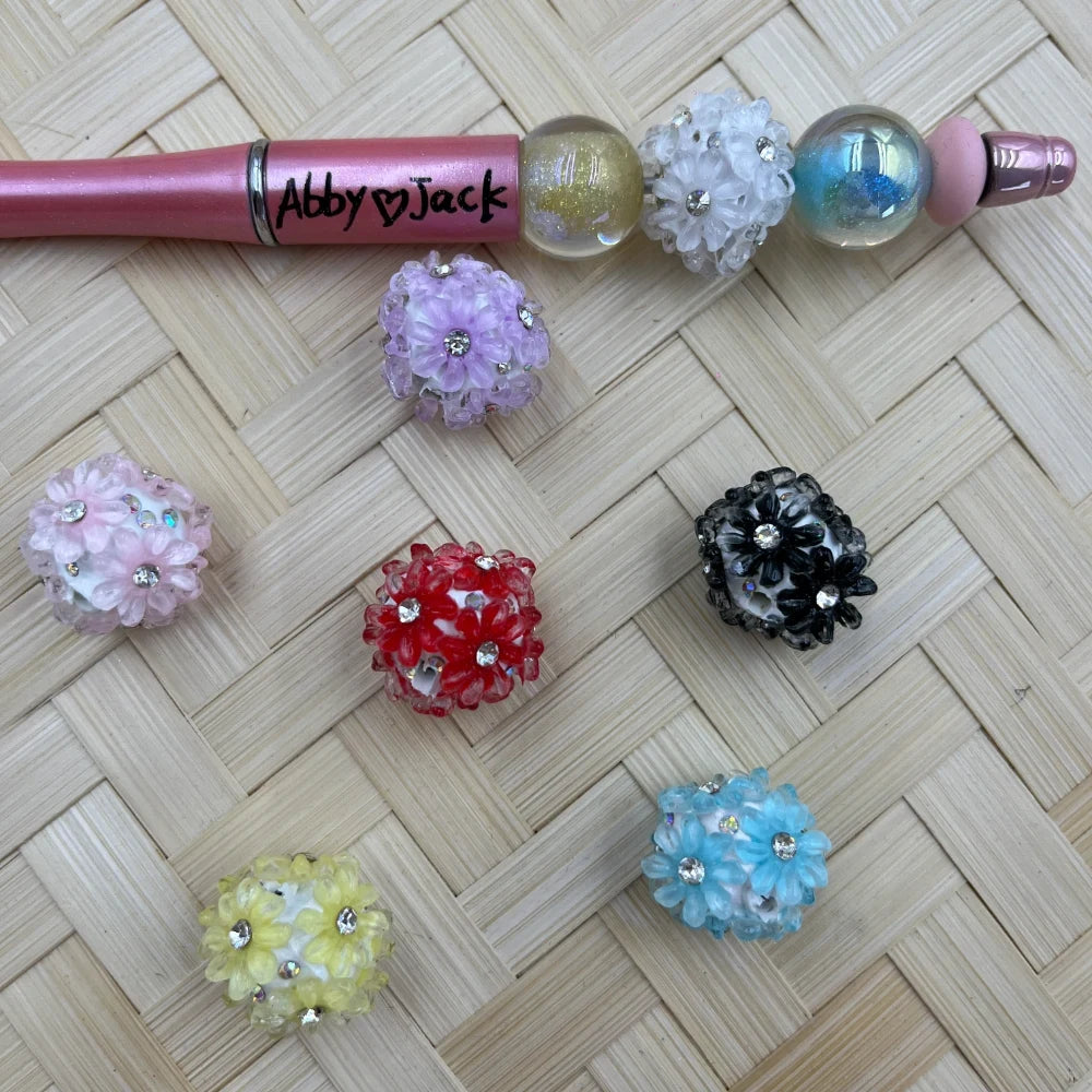 Assorted rhinestone flower cluster fancy beads in pink, purple, red, black, yellow, blue, and white displayed on a woven surface next to a pink beadable pen labeled Abby Jack.