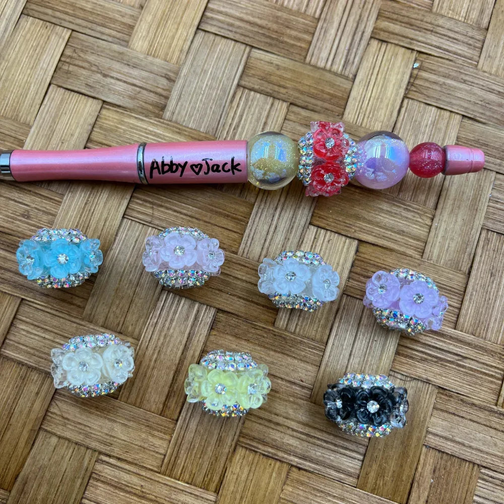 Assorted rhinestone flower cluster fancy beads in blue, pink, white, purple, yellow, clear, and black displayed on a wicker surface next to a pink beadable pen labeled Abby Jack.