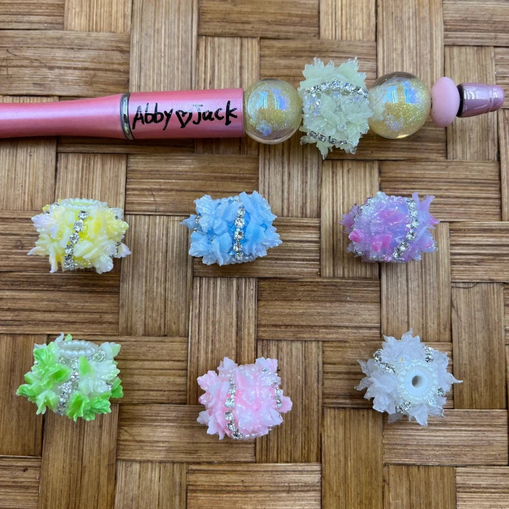 Assorted pastel rhinestone flower cluster fancy beads in yellow, blue, purple, pink, green, and white displayed on a woven surface next to a pink beadable pen labeled Abby and Jack
