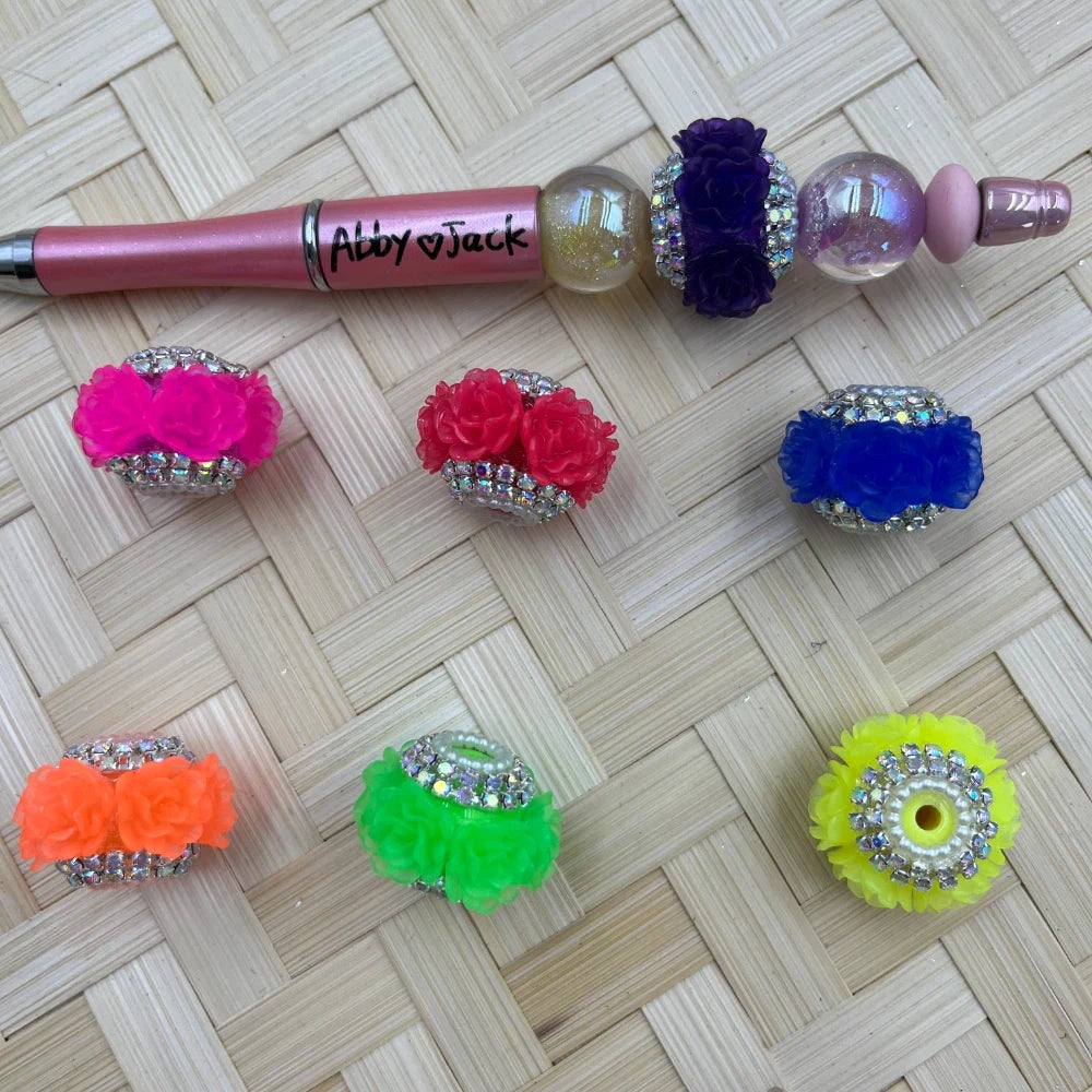 Colorful rhinestone flower fancy beads in pink, red, blue, orange, green, yellow, and purple displayed on a woven surface next to a custom beadable pen labeled Abby and Jack.