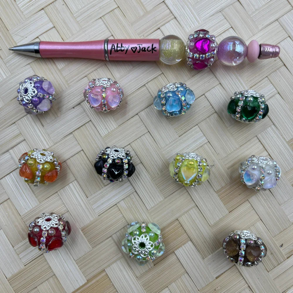 Assorted rhinestone flower petal fancy beads in multiple colors including purple, pink, blue, green, black, yellow, red, and brown displayed on woven surface next to a decorated beadable pen labeled Abby Jack.