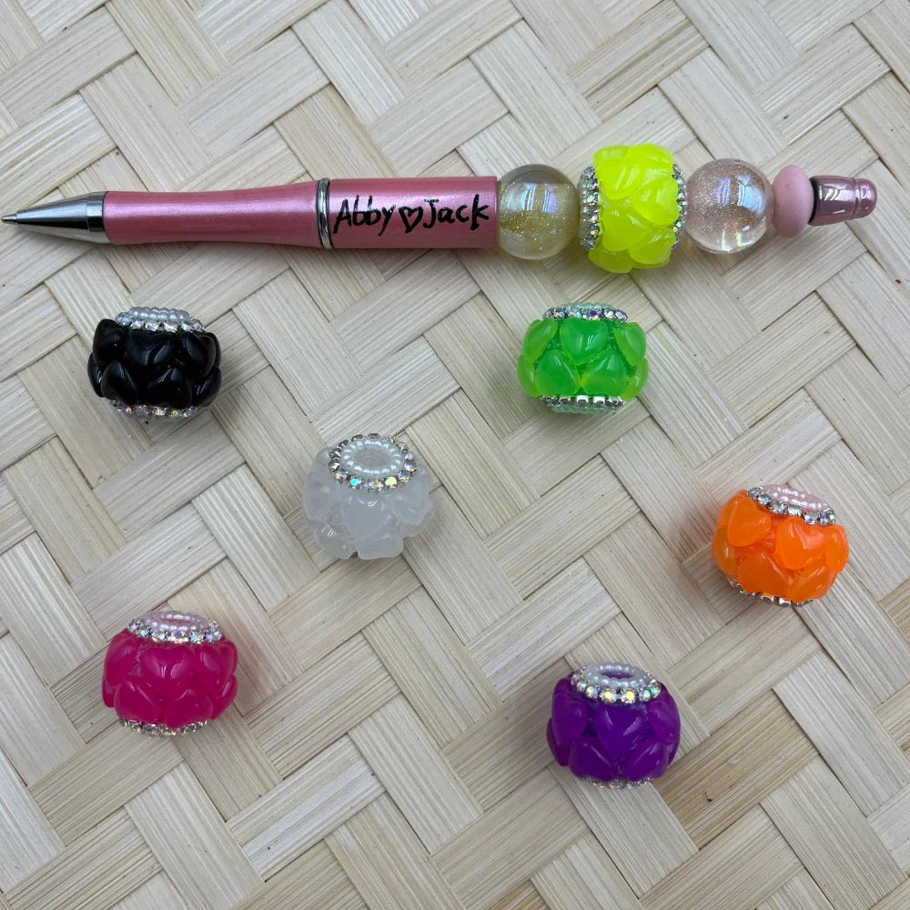Colorful rhinestone-trimmed flower resin fancy beads in black, white, pink, green, yellow, orange, and purple displayed with a pink beadable pen on a woven surface.