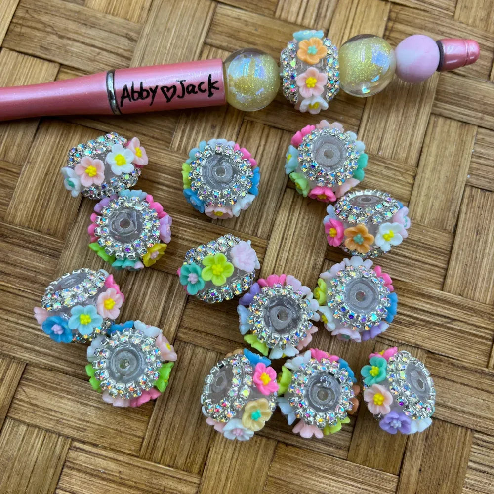Colorful pastel polymer flower rhinestone rondelle fancy beads displayed on wicker surface next to a decorated beadable pen branded Abby Jack, showing multicolor floral embellishments and sparkling crystal rhinestone borders.